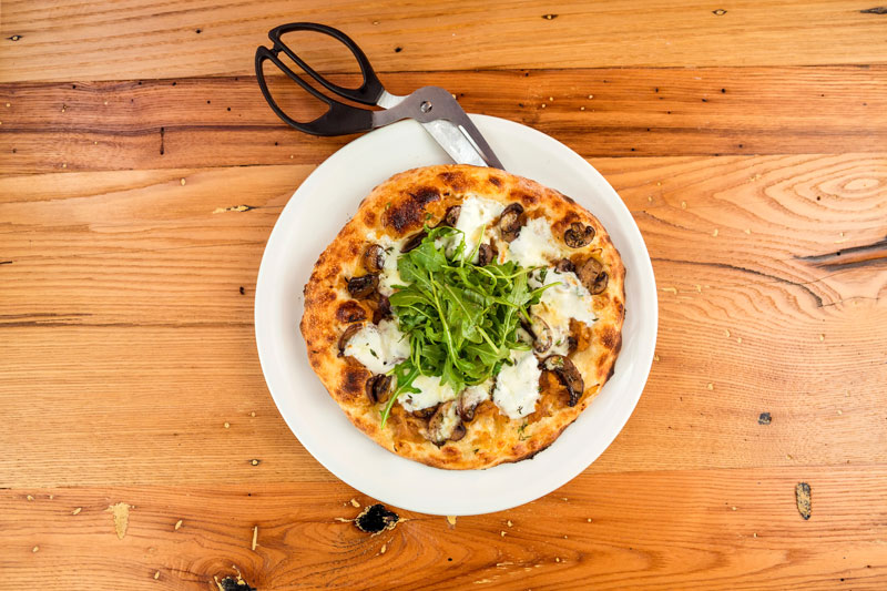 overhead view of white pizza topped with mushrooms and greens with crispy crust on a light wooden table